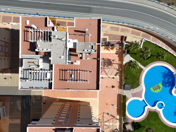 Aerial view of a rooftop complex with red tiles, white railings, and a curved road at the top; a blue pool and palm trees on the right.