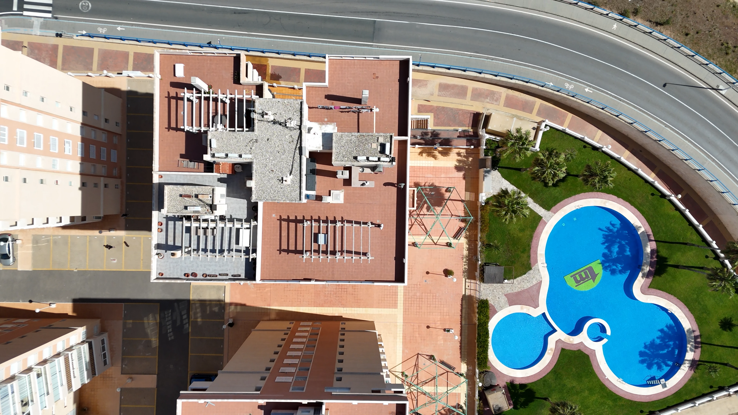 Aerial view of a rooftop complex with red tiles, white railings, and a curved road at the top; a blue pool and palm trees on the right.