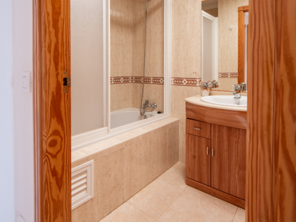 Bathroom interior with a glass-enclosed shower and beige tile walls, a white sink on a wooden vanity, and a mirror reflecting the shower area (neutral tones).