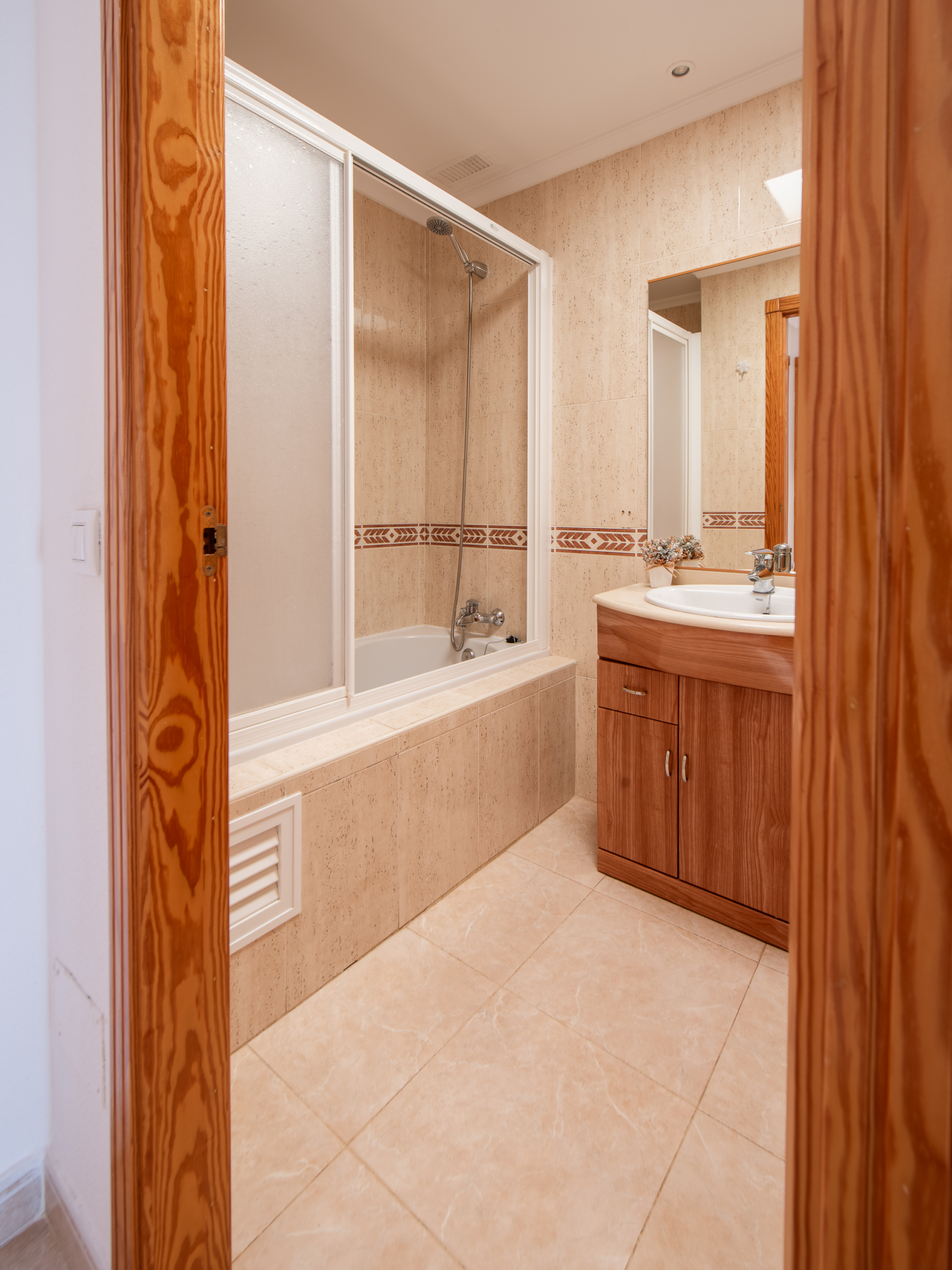 Bathroom interior with a glass-enclosed shower and beige tile walls, a white sink on a wooden vanity, and a mirror reflecting the shower area (neutral tones).