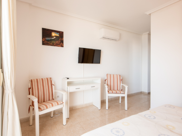 Bright hotel room with a white desk and two striped-foam chairs facing a wall-mounted TV, an air conditioner above, and a glimpse of a bed in the foreground.