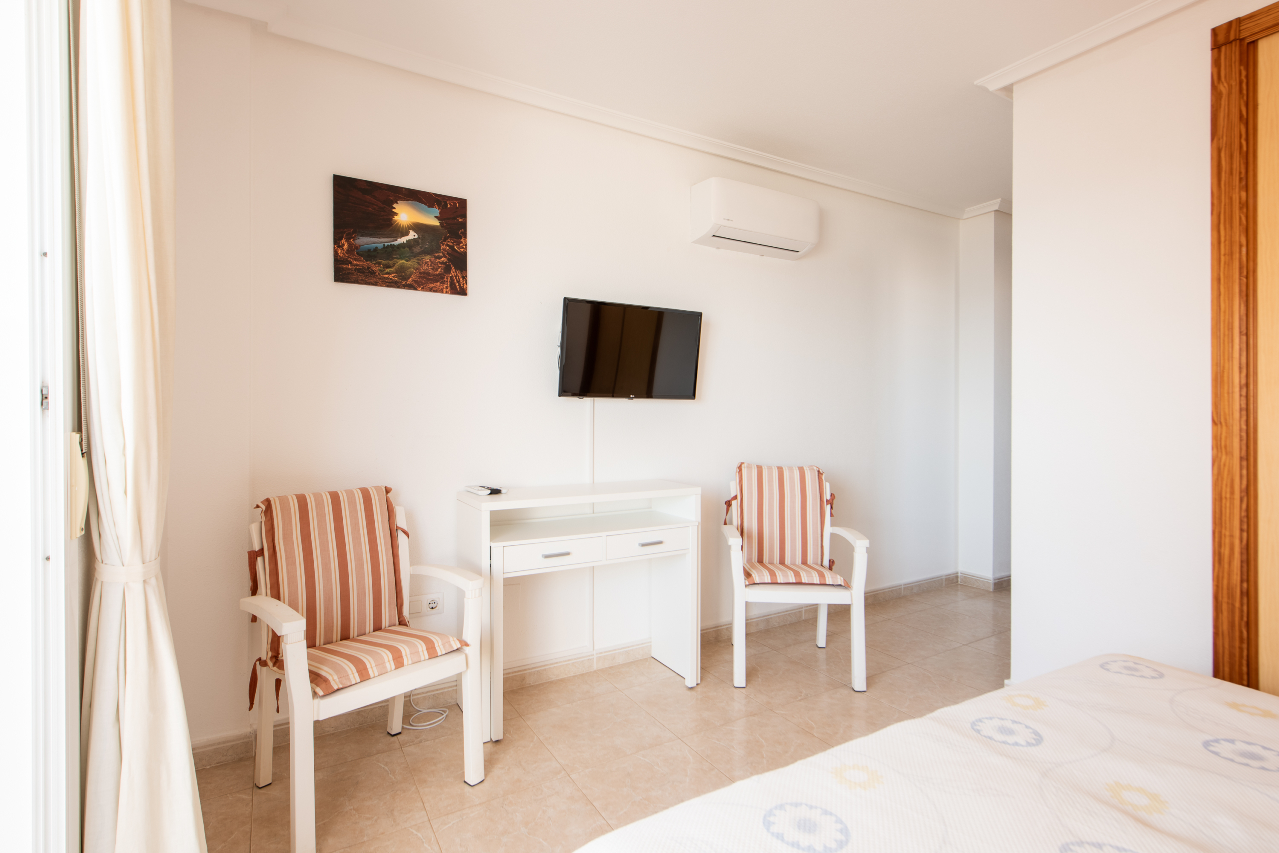 Bright hotel room with a white desk and two striped-foam chairs facing a wall-mounted TV, an air conditioner above, and a glimpse of a bed in the foreground.