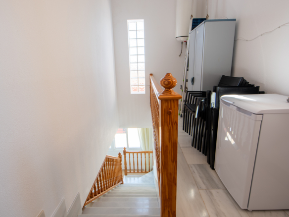 Bright stairwell with a wooden banister, white walls, and a laundry area with a cabinet and stacked chairs to the side.