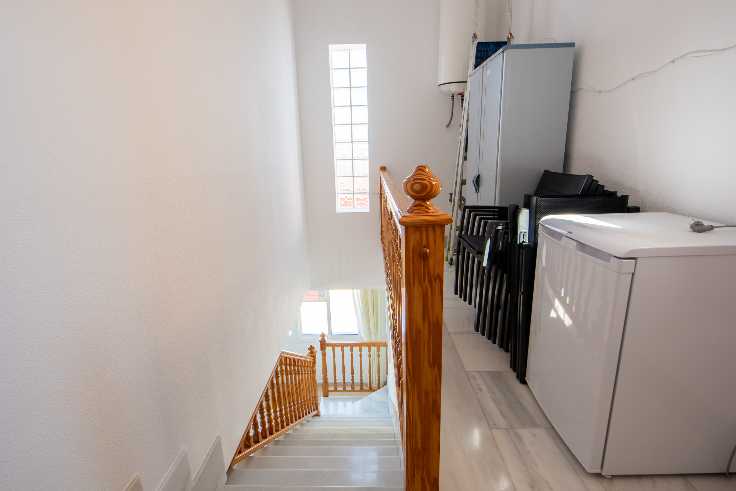 Bright stairwell with a wooden banister, white walls, and a laundry area with a cabinet and stacked chairs to the side.