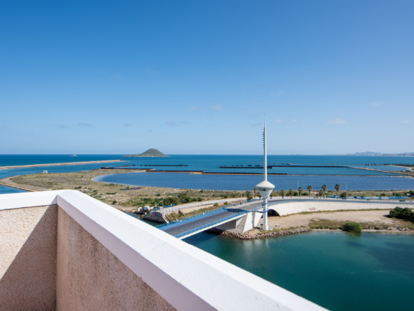 Sunny coastal view with calm blue sea, small island on the horizon, and a pale pink balcony wall in the foreground.
