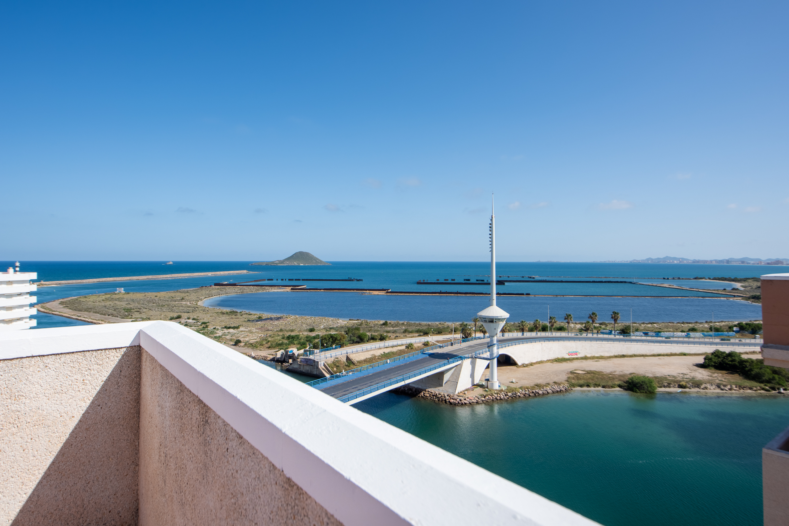 Sunny coastal view with calm blue sea, small island on the horizon, and a pale pink balcony wall in the foreground.