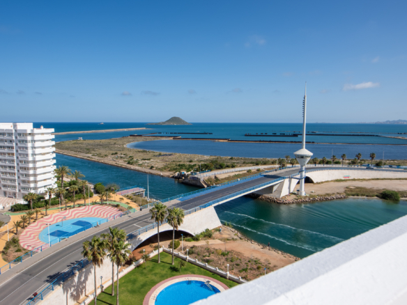 Coastal cityscape with white apartment buildings, palm trees, a blue bay, a tall observation tower, and a circular pool in the foreground under a clear sky.