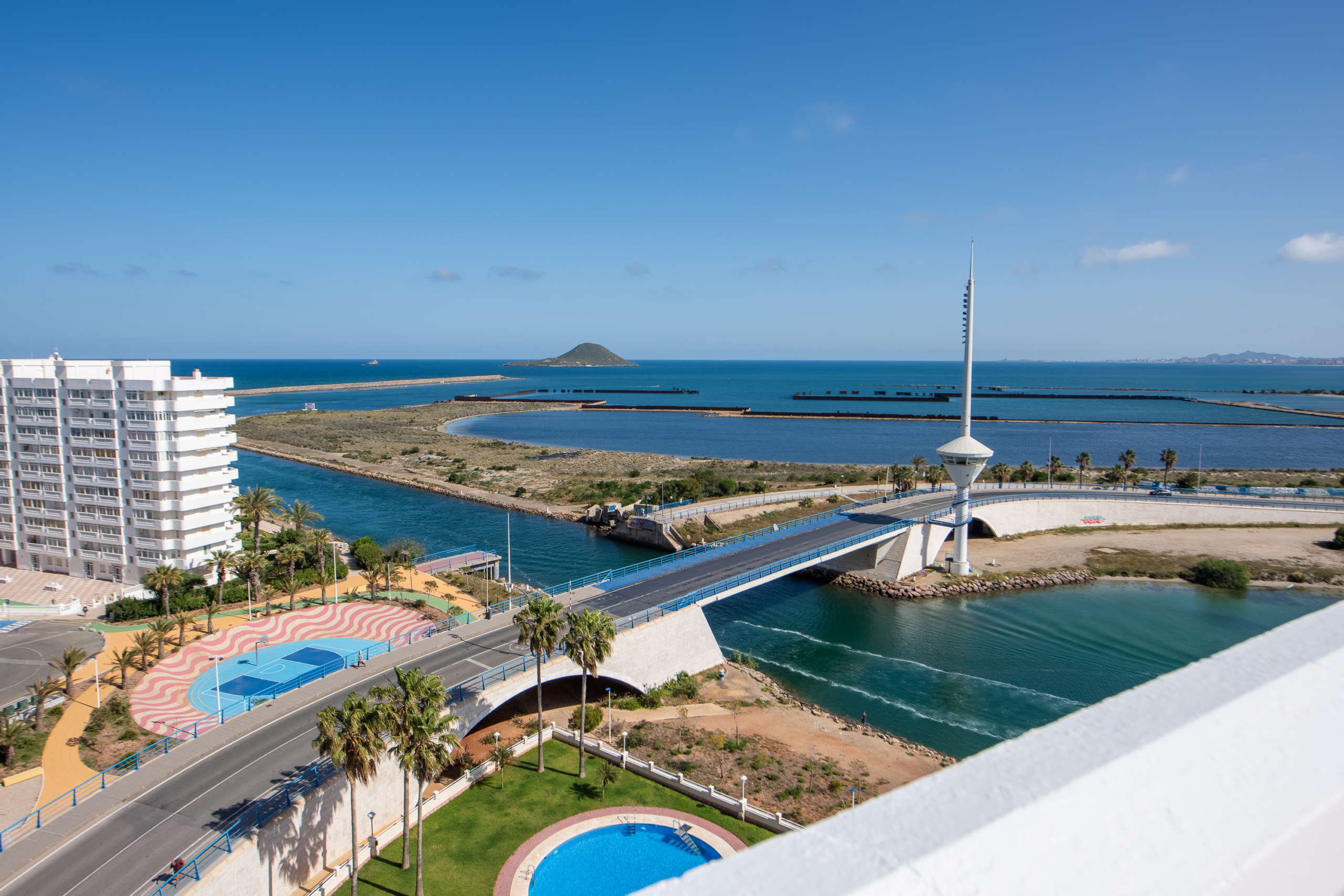 Coastal cityscape with white apartment buildings, palm trees, a blue bay, a tall observation tower, and a circular pool in the foreground under a clear sky.