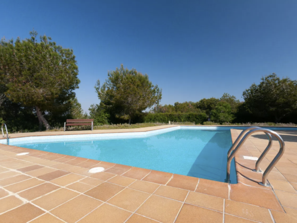 Outdoor swimming pool surrounded by tan tile deck, metal ladders, and a wooden bench under a clear blue sky. Nearby trees provide greenery.