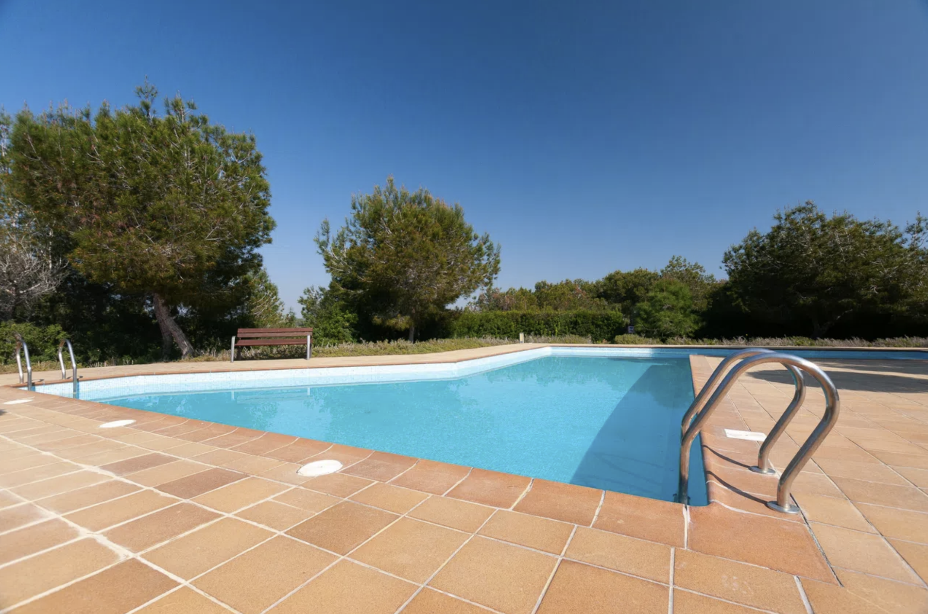 Outdoor swimming pool surrounded by tan tile deck, metal ladders, and a wooden bench under a clear blue sky. Nearby trees provide greenery.