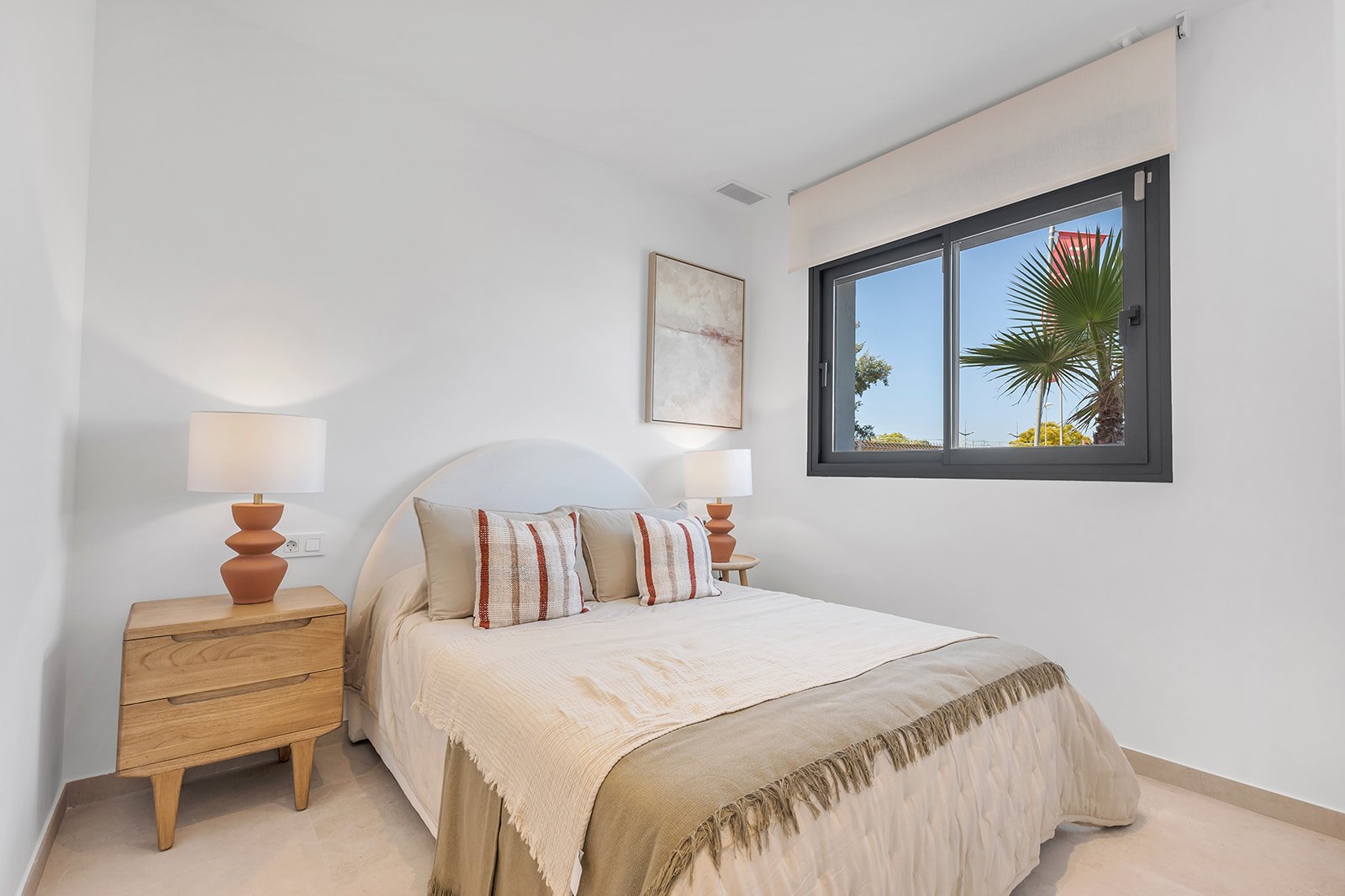 Een slaapkamer met een bed, nachtkastje en raam met uitzicht op een palmboom.