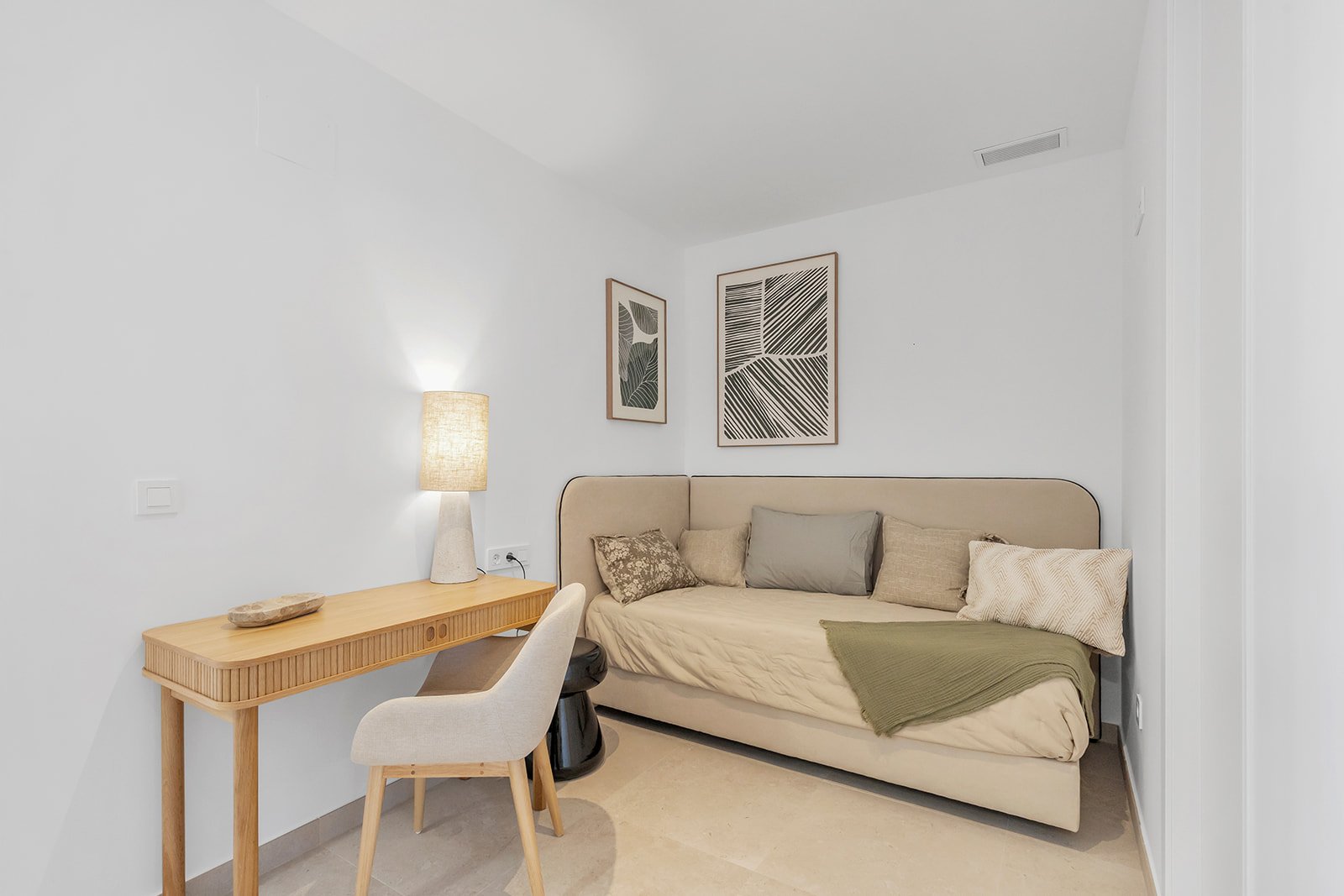 Een ruimte met een bedbank, bureau met stoel en een lamp, plus wanddecoraties.