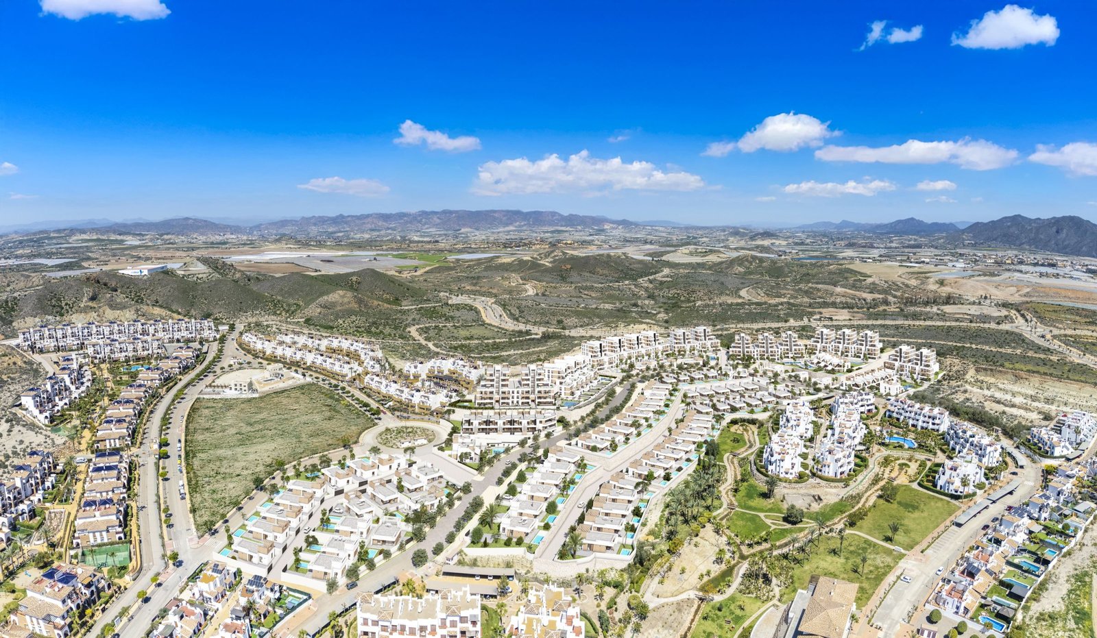 Luchtfoto van een woonwijk met woningen, wegen en groene zones, met heuvels op de achtergrond.
