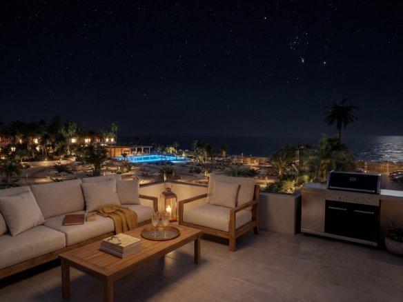 Terras met zithoek en buitenkeuken, met op de achtergrond een verlicht zwembad en de zee bij nacht.